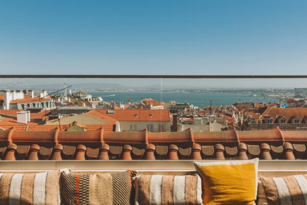 Um terraço no último piso com almofadas às riscas e amarelas tem vista para os telhados vermelhos, uma paisagem urbana e uma massa de água azul sob um céu limpo.