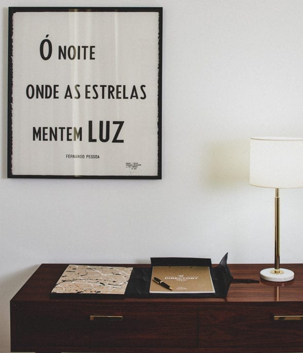 A minimal interior with a framed Portuguese quote on the wall, a table lamp with a white shade, and a wooden sideboard holding an open folder, a marble-patterned notebook, and a brown covered book.