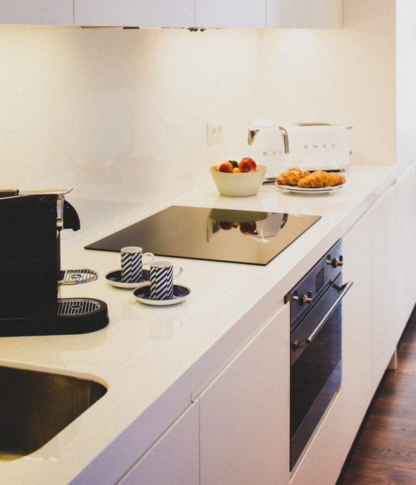 Une cuisine moderne avec un plan de travail blanc, une cuisinière électrique, un évier, une machine à café noire, deux tasses à café assorties, un bol de fruits, un grille-pain, une bouilloire et une assiette de croissants.