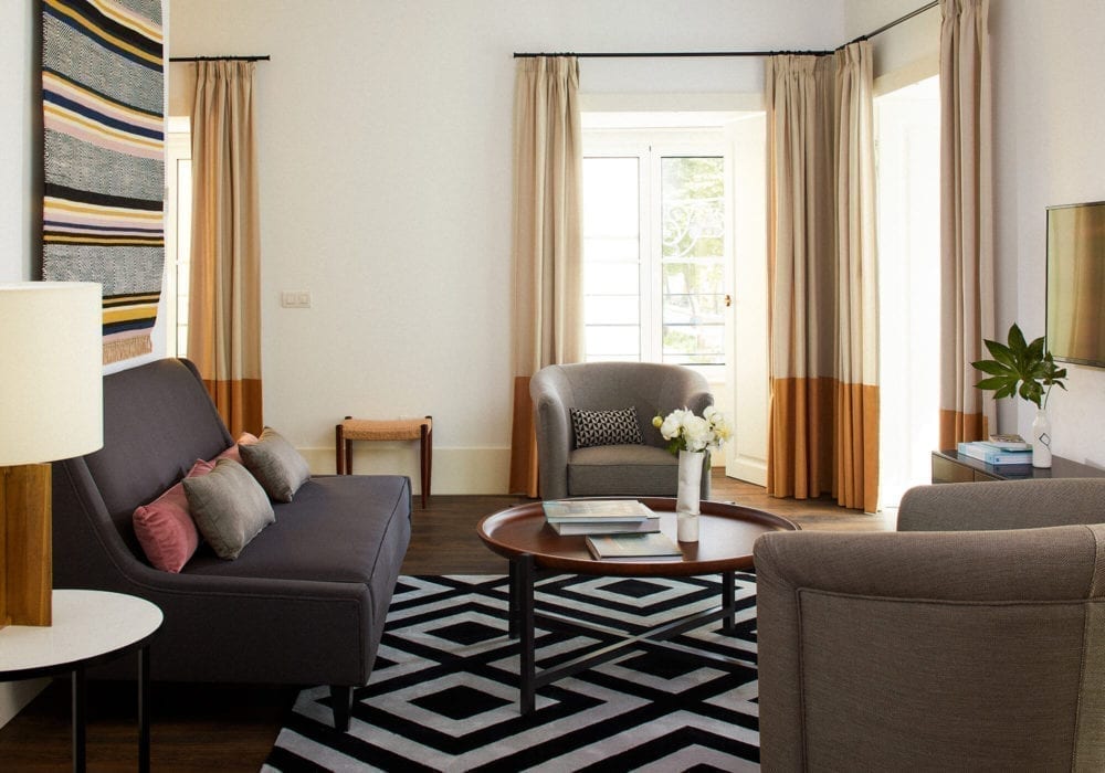 Salon moderne avec un canapé foncé, deux fauteuils gris, un tapis géométrique noir et blanc, une table basse ronde en bois avec des fleurs et des livres, de grandes fenêtres avec des rideaux beiges et une télévision murale.