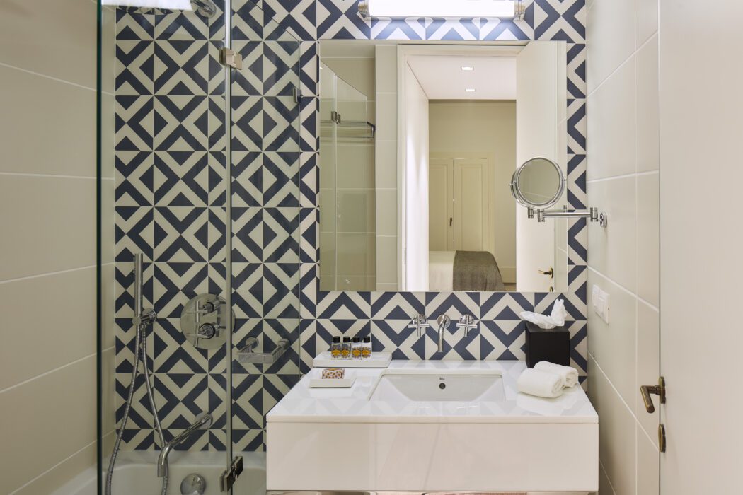 Modern bathroom with geometric blue-and-white tile accent wall, glass shower, rectangular white sink, mirror, towel, and toiletries neatly arranged on the counter. Bright lighting and clean, minimalist decor.