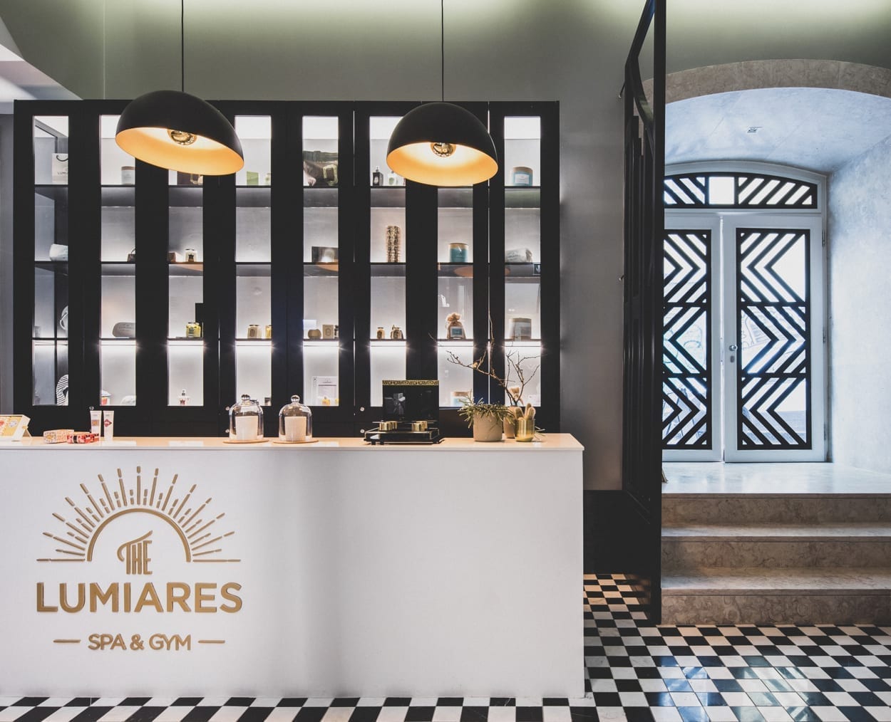 Reception desk of The Lumiares Spa & Gym with a modern, stylish interior. Black and white checkered floor, illuminated shelving with products, two hanging lights, and decorative black-and-white doors in the background.