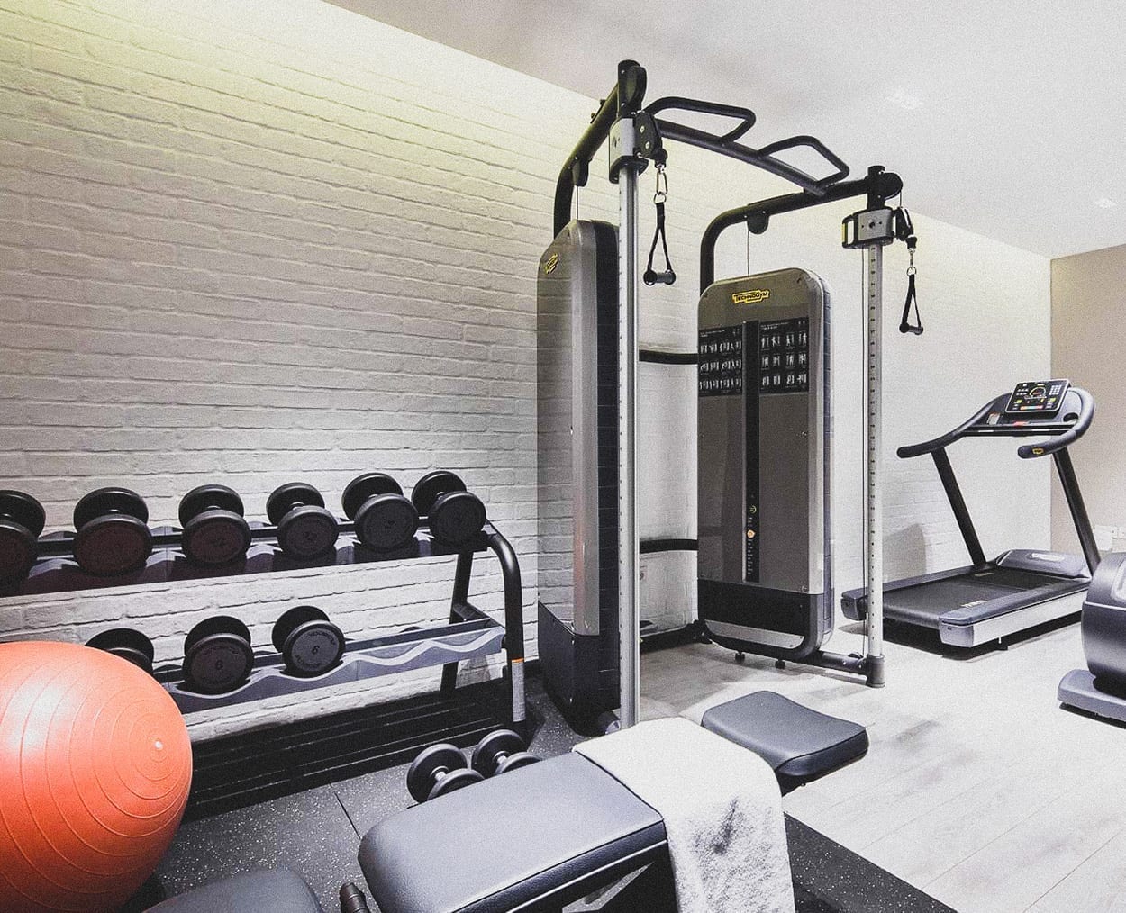 A modern gym room with a rack of dumbbells, a red exercise ball, a cable machine, a treadmill, and a weight bench with a towel on it, all set against a white brick wall.