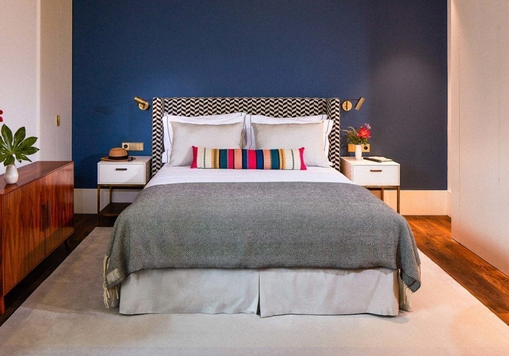 A modern bedroom with a bed centered against a dark blue accent wall, colorful patterned headboard, gray blanket, striped pillow, white nightstands with lamps, and wood furniture on a light rug.