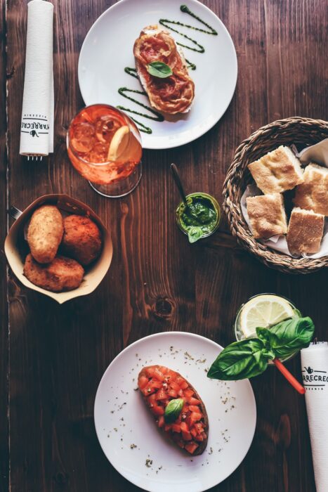 Vista aérea de una comida italiana en una mesa de madera, con bruschetta, cubos de pan en una cesta, bocadillos fritos, una salsa de hierbas verdes y dos bebidas, una con una rodaja de naranja y otra con una guarnición de lima y albahaca.