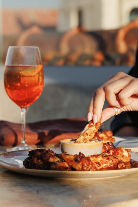 Uma mão mergulha um pedaço de carne grelhada ou de pão num molho cremoso num prato, com um copo de bebida cor de laranja ao lado, numa mesa ao ar livre, sob a luz quente do sol.