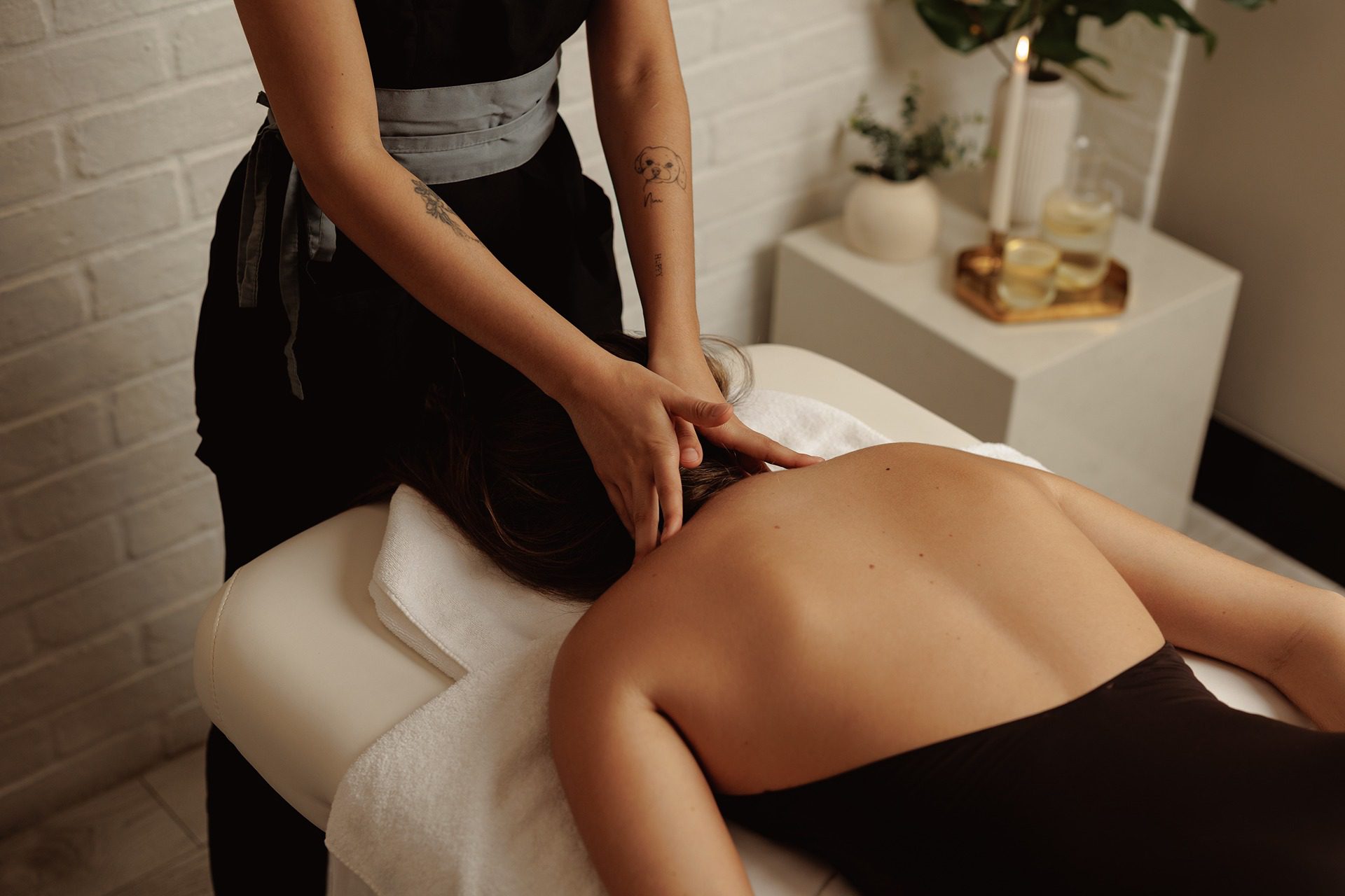 A person receiving a neck and shoulder massage while lying face down on a massage table, with a massage therapist standing beside them in a serene, softly lit room.