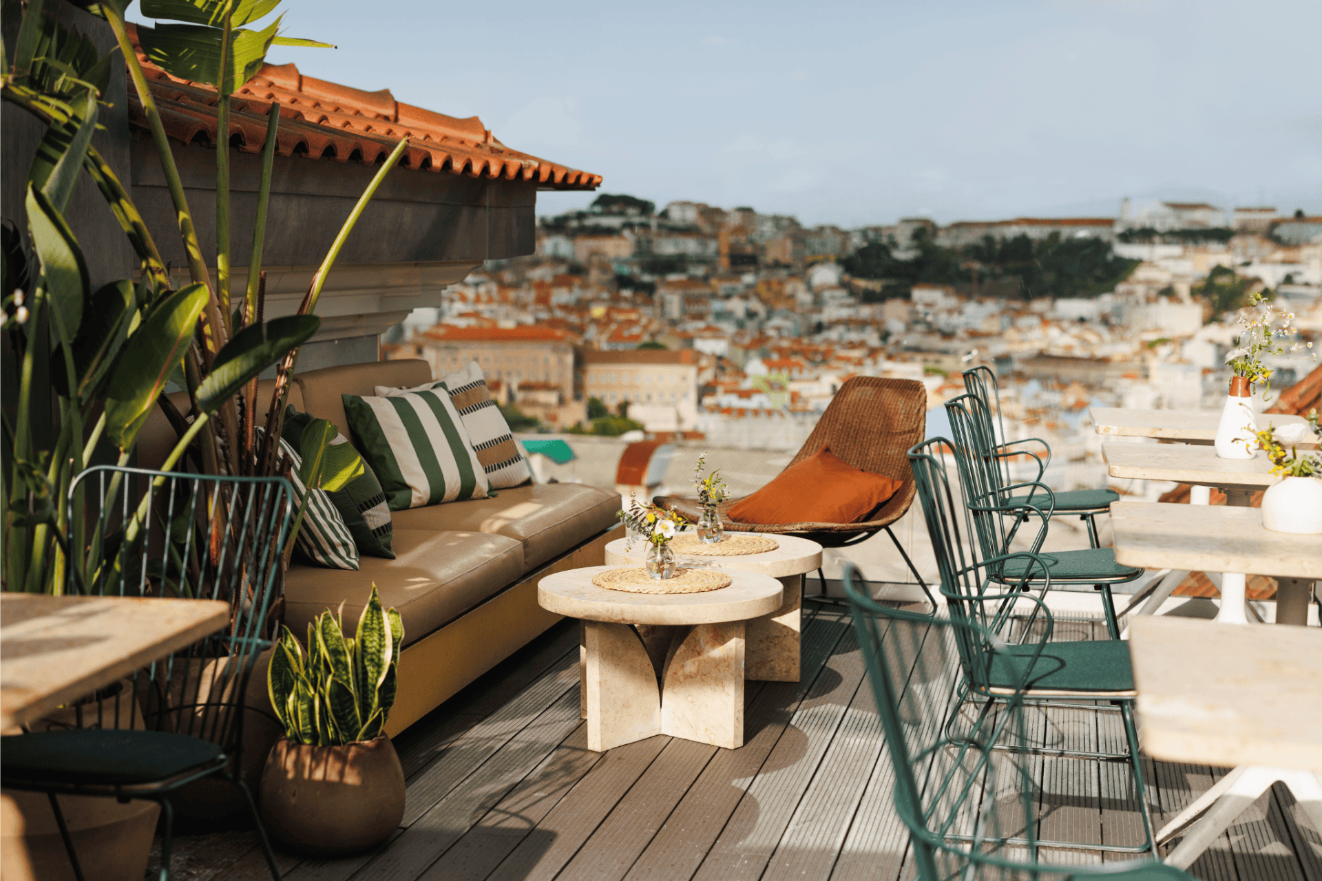 A rooftop terrace with a cushioned sofa, lounge chair, small tables, and green metal chairs overlooks a cityscape with buildings and hills in the distance. Potted plants decorate the cozy outdoor seating area.