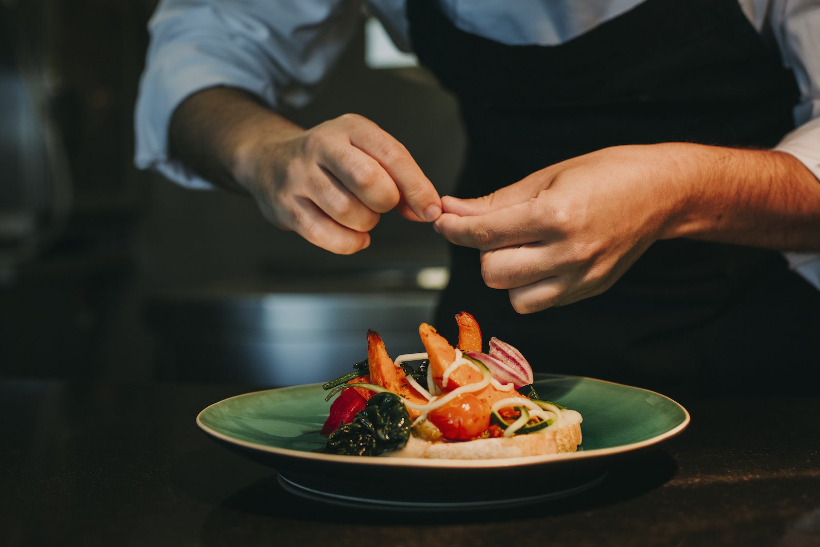 A chef garnishes a plated dish with shrimp, vegetables, and greens, carefully arranging ingredients on a green-rimmed plate in a dimly lit kitchen.
