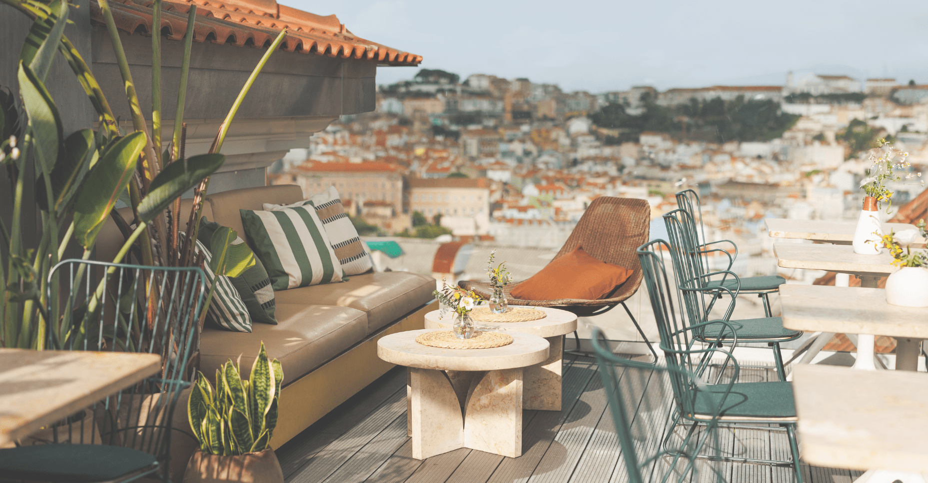 Rooftop terrace with a beige sofa, striped cushions, small tables, plants, and metal chairs overlooking a cityscape with hillside buildings in the background under a clear sky.