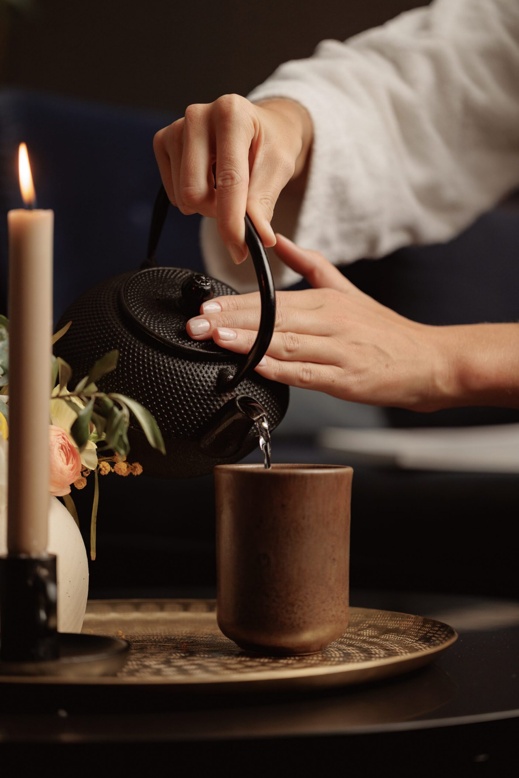 A person pours tea from a textured black teapot into a brown ceramic cup on a woven tray beside a lit candle and a vase with flowers.