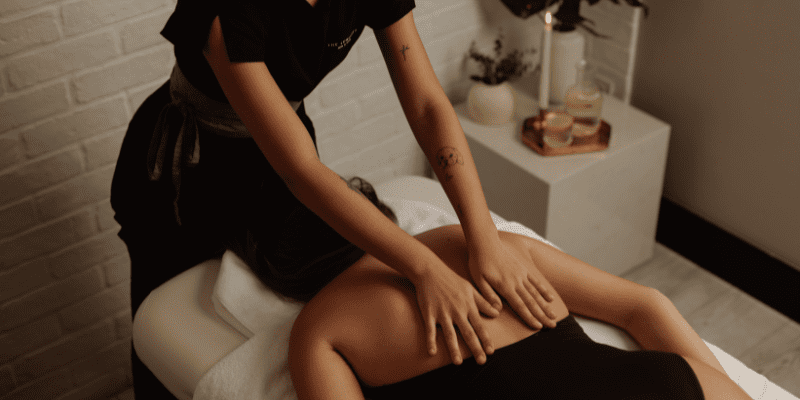 A person receives a back massage while lying face down on a massage table in a softly lit room with minimal decor. The massage therapist is standing beside the table, using both hands to massage the person’s back.