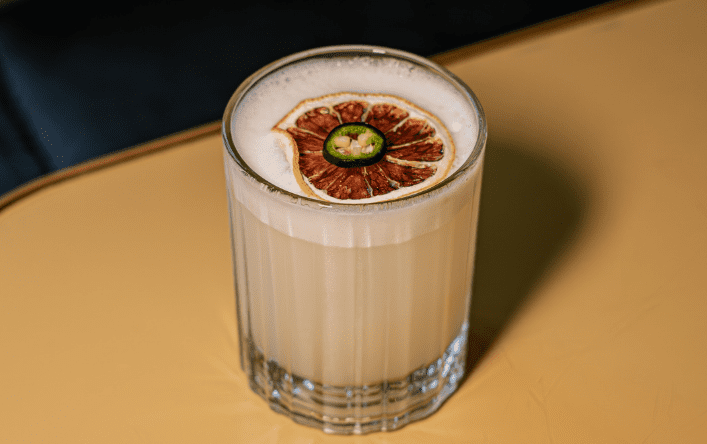 A creamy cocktail in a clear glass, topped with a dried citrus slice and a small green chilli pepper, sits on a light tan surface.