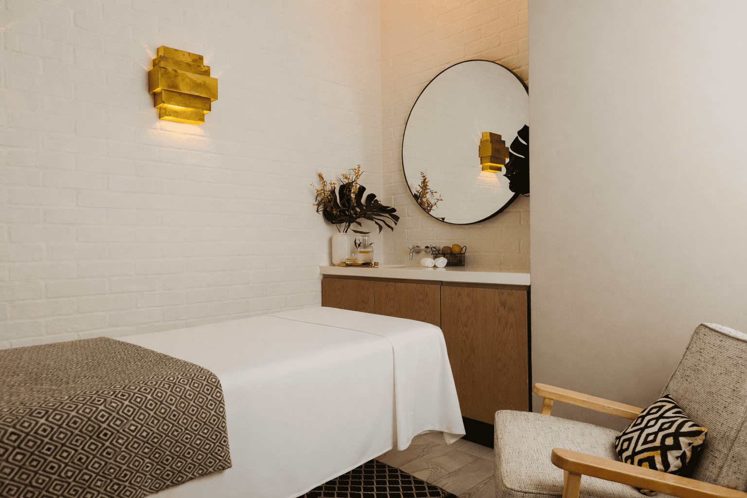 A cosy spa room with a massage table covered in white sheets, a patterned throw, a wooden cupboard with a round mirror above it, modern gold lighting, and a beige armchair with a decorative cushion.