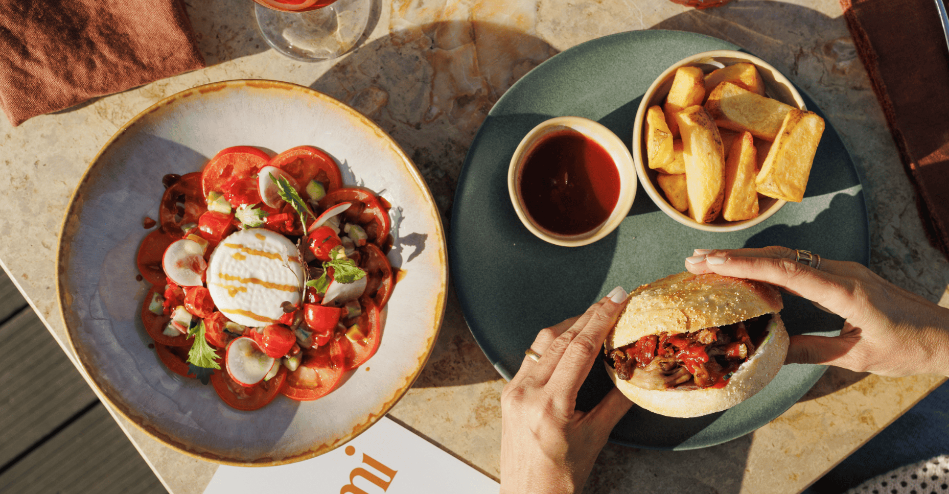 A person holds a sandwich with roasted vegetables. On the table are thick-cut chips with tomato sauce and a salad with tomatoes, radishes, herbs, and cheese on a marble surface.