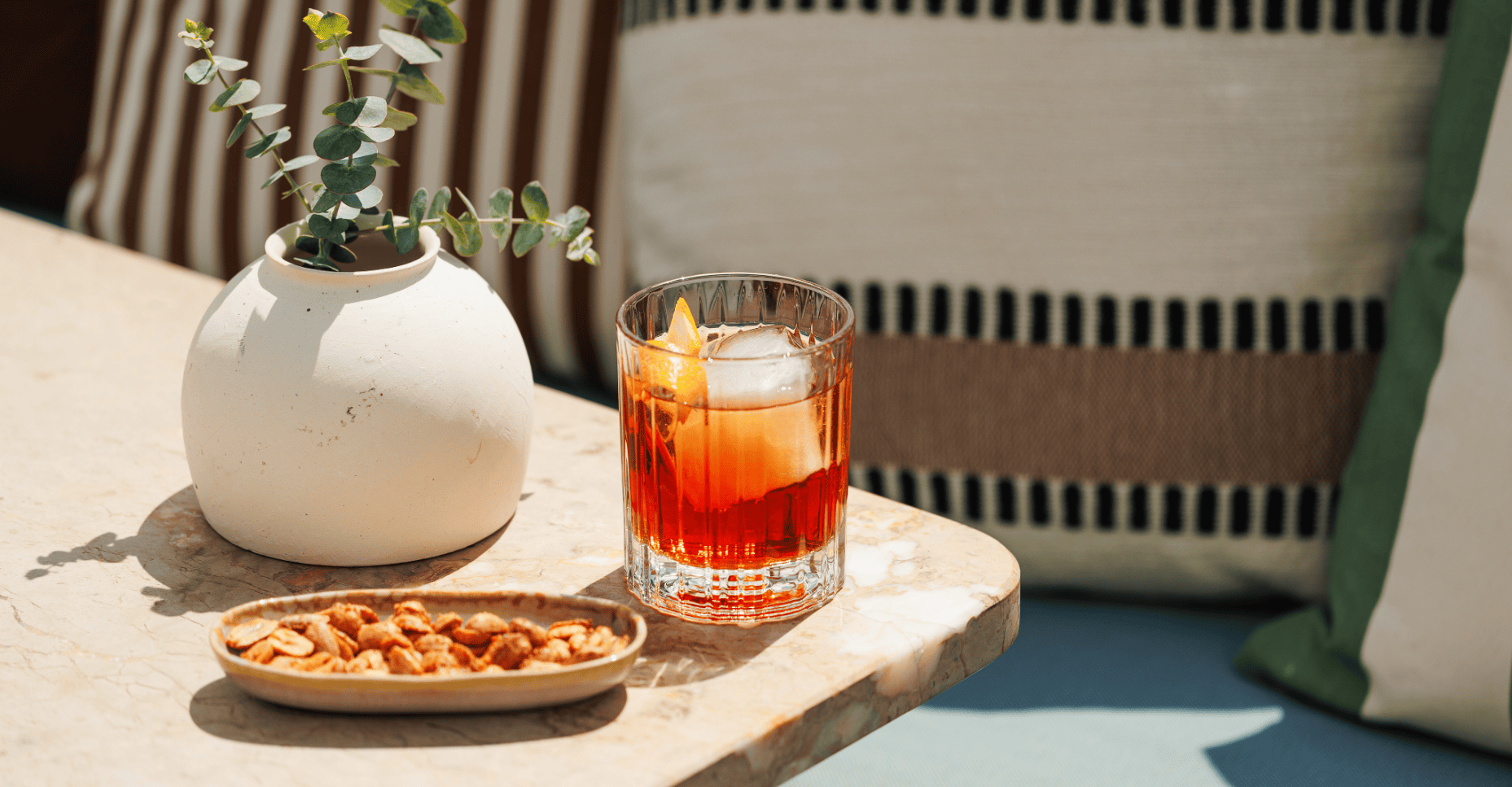 A glass of iced red cocktail with an orange slice, a small dish of snacks, and a round vase with greenery on a sunlit table, with striped cushions in the background.