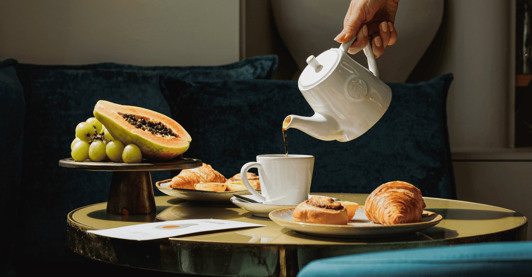 A hand pours tea from a white teapot into a cup on a table with pastries, grapes, and half a papaya on a stand, set against a dark green sofa.