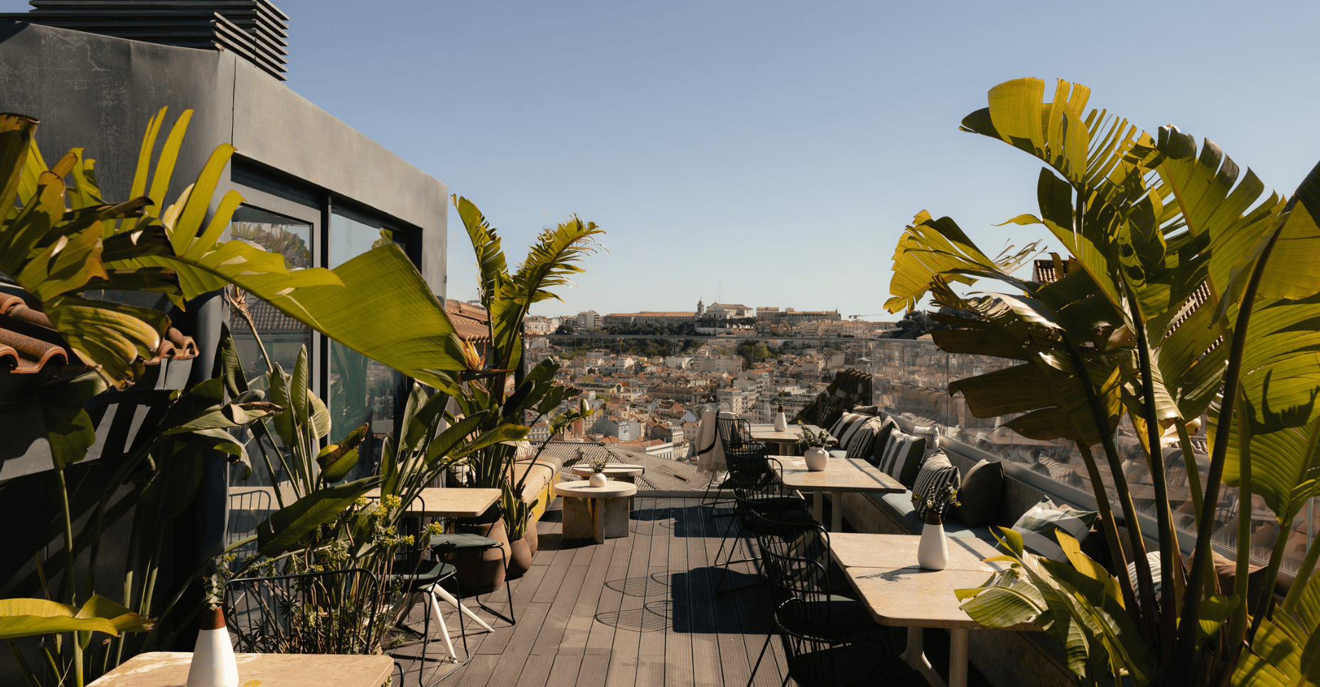 A rooftop terrace with tables, chairs, and potted plants overlooks a cityscape under a clear blue sky.
